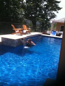 A child splashes in a bright blue pool while being held by an adult beside wooden chairs.