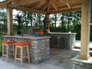Outdoor kitchen featuring a stone bar, wooden stools, grill, and greenery in the background.