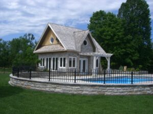 Charming stone house with a gable roof beside a circular pool and lush green lawn.