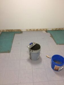 Gray and blue buckets on tiled floor with partially laid green tiles. Walls are bare.