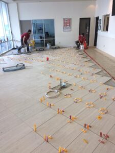 Two workers are installing tile flooring with spacers, tools, and equipment visible in the room.