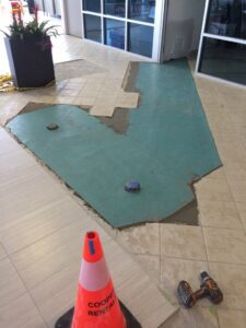 Floor tiles are being replaced, revealing green backing and a warning cone nearby.