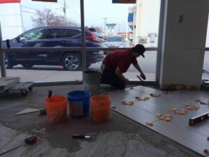 A worker lays tiles on the floor of a commercial space, with buckets and tools nearby.