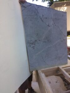 A person holds a large gray marble slab against a white surface outdoors.