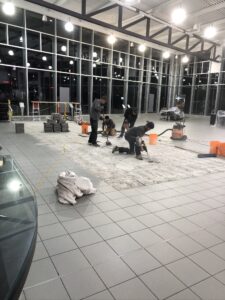 Workers lay new tiles on a floor, with stacks of tiles and construction tools visible.