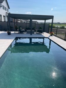 Modern patio with a pool, lounge seating, and a shaded area under a structure with a clear sky above.
