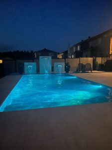 Illuminated pool at night with three water features and surrounding modern homes.